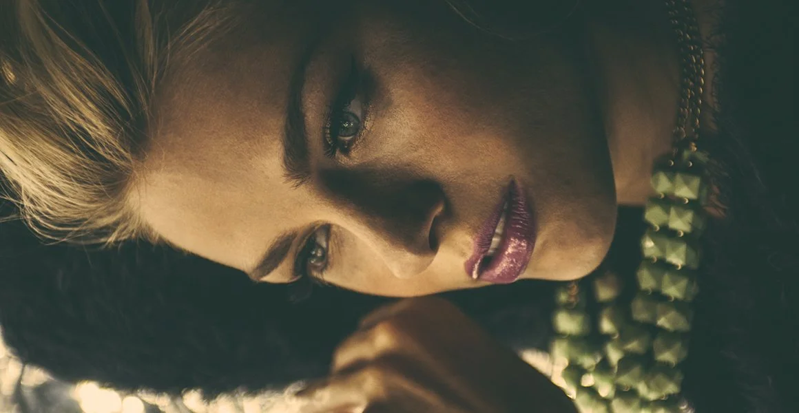 Close-up of a woman with blonde and black hair, looking downward with a contemplative expression, wearing a black top and layered necklace.