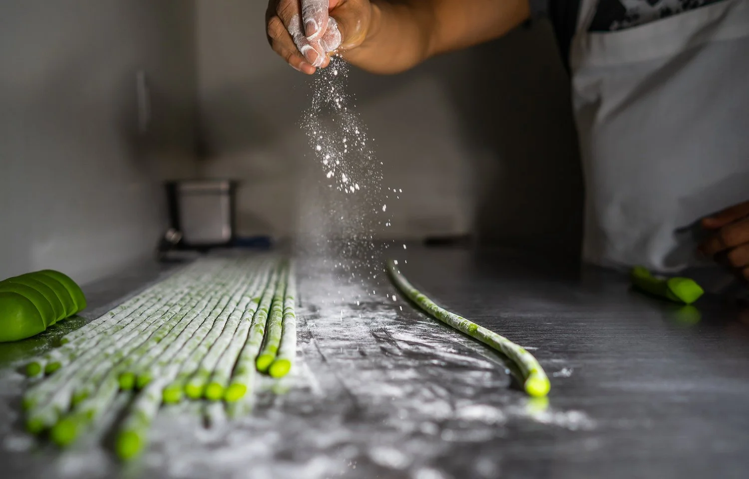 A chef in a kitchen sprinkling flour onto rolled green dough sticks on a countertop.
