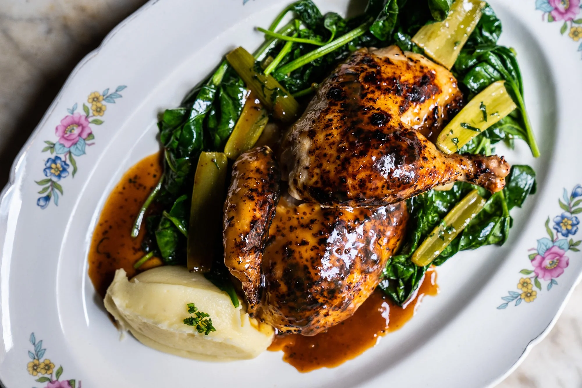 A plate with roasted chicken covered in sauce, mashed potatoes topped with chives, and sautéed greens with celery stalks.