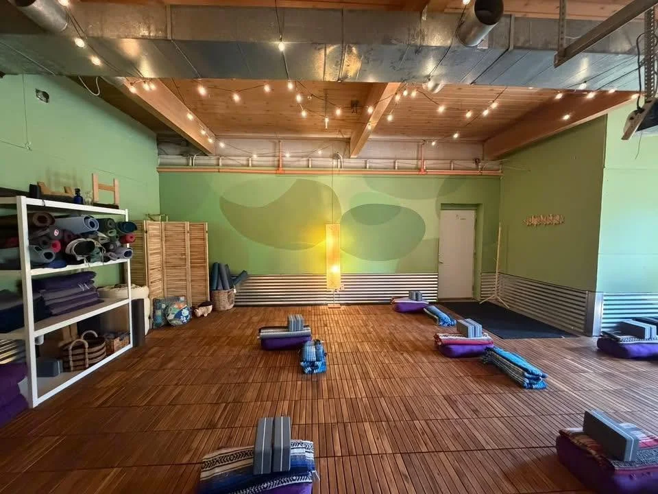 Image is of the DTYC studio. There are green walls, wood floors, and yoga props are set up across the room, prepped for a class. There are floor lamps and patio string lights strung across the ceiling.