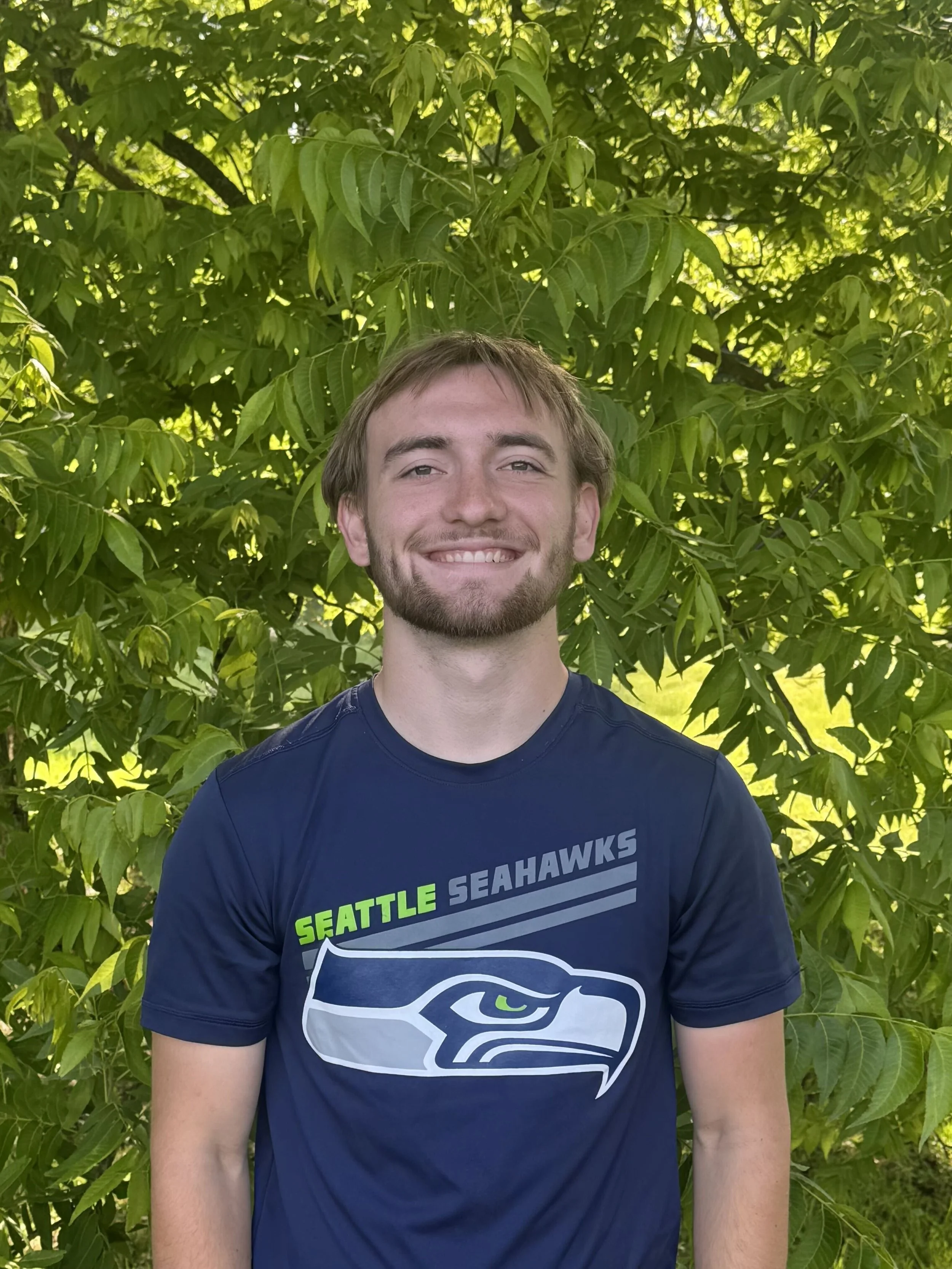 A white man with short, light brown hair and short facial hair wears a Seattle Seahawks t-shirt in front of a foliage filled background