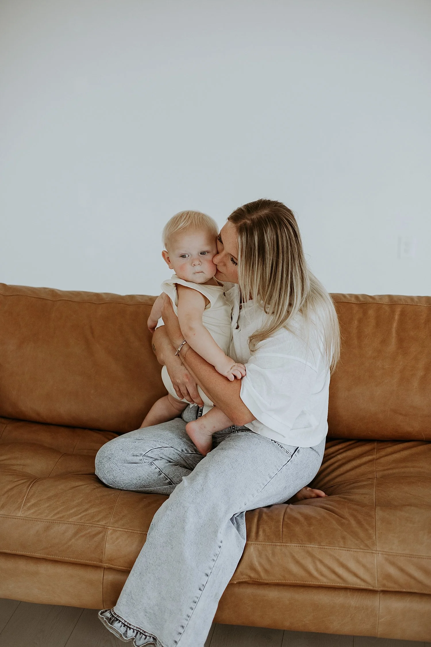  blonde mom in jeans snuggles baby on leather couch by Round Rock Family Photographer 