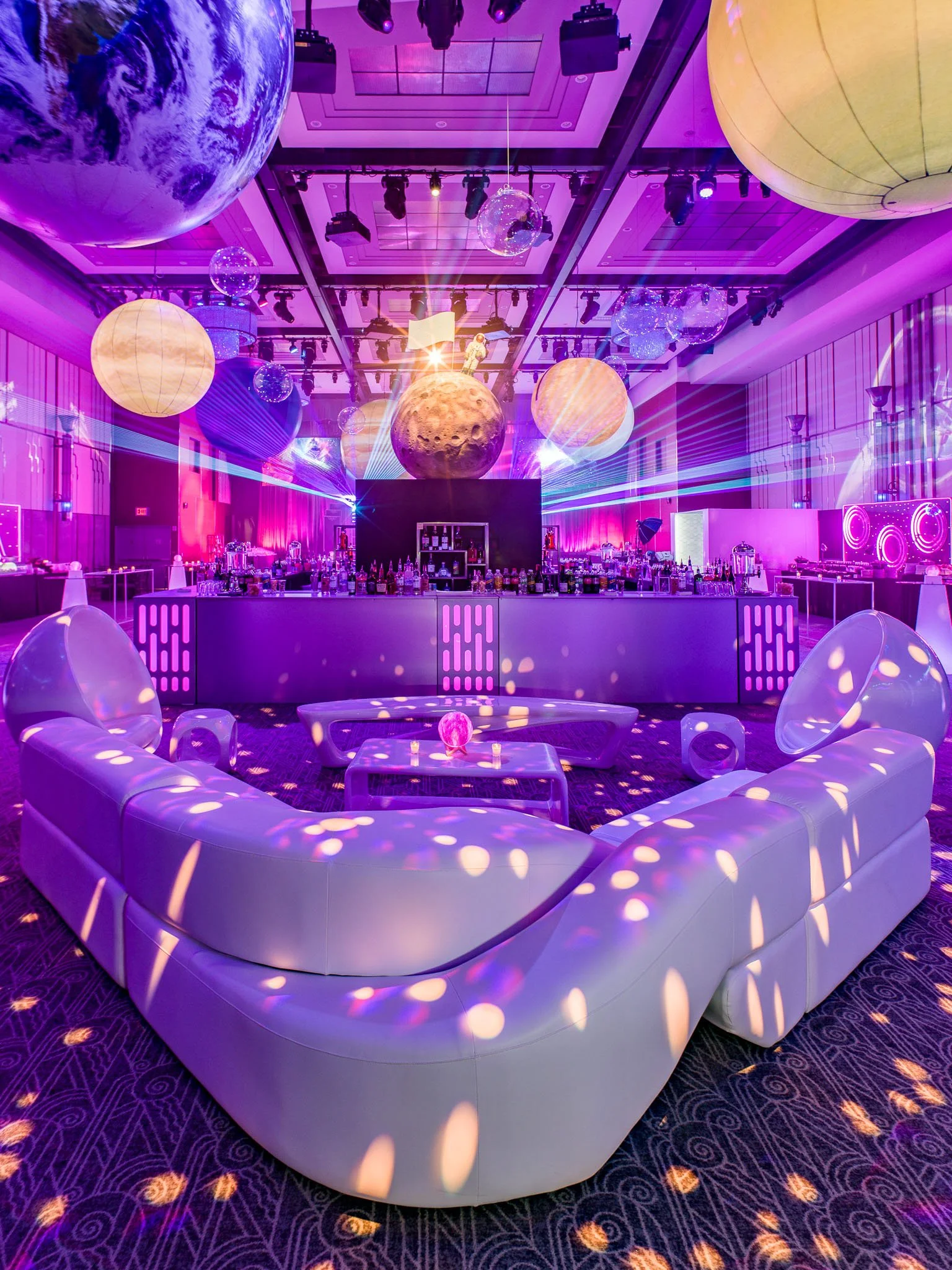A futuristic nightclub with space-themed decorations, including moon and planet models hanging from the ceiling, colorful laser lights, and a bar area, with a curved white sofa and small tables in the foreground.