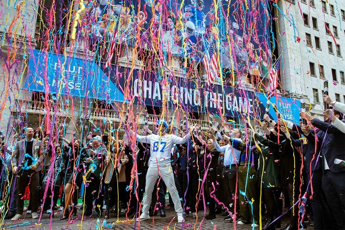 Celebration scene on Wall Street with a large crowd, colorful confetti, and streamers, people taking photos, a man with arms raised, large screens displaying images and text about the NYSE and a changing game, with a city building in the background.