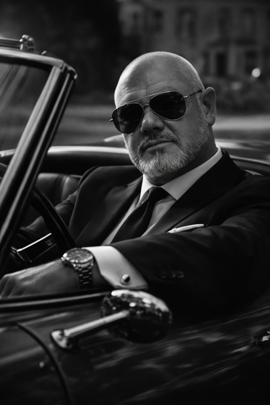 Scott Morgan, the man behind Crown Imagery. 15 years of hard work show him sitting in a classic sports car in a tailored suit and Ray-Ban sunglasses. Shaved head and trimmed beard