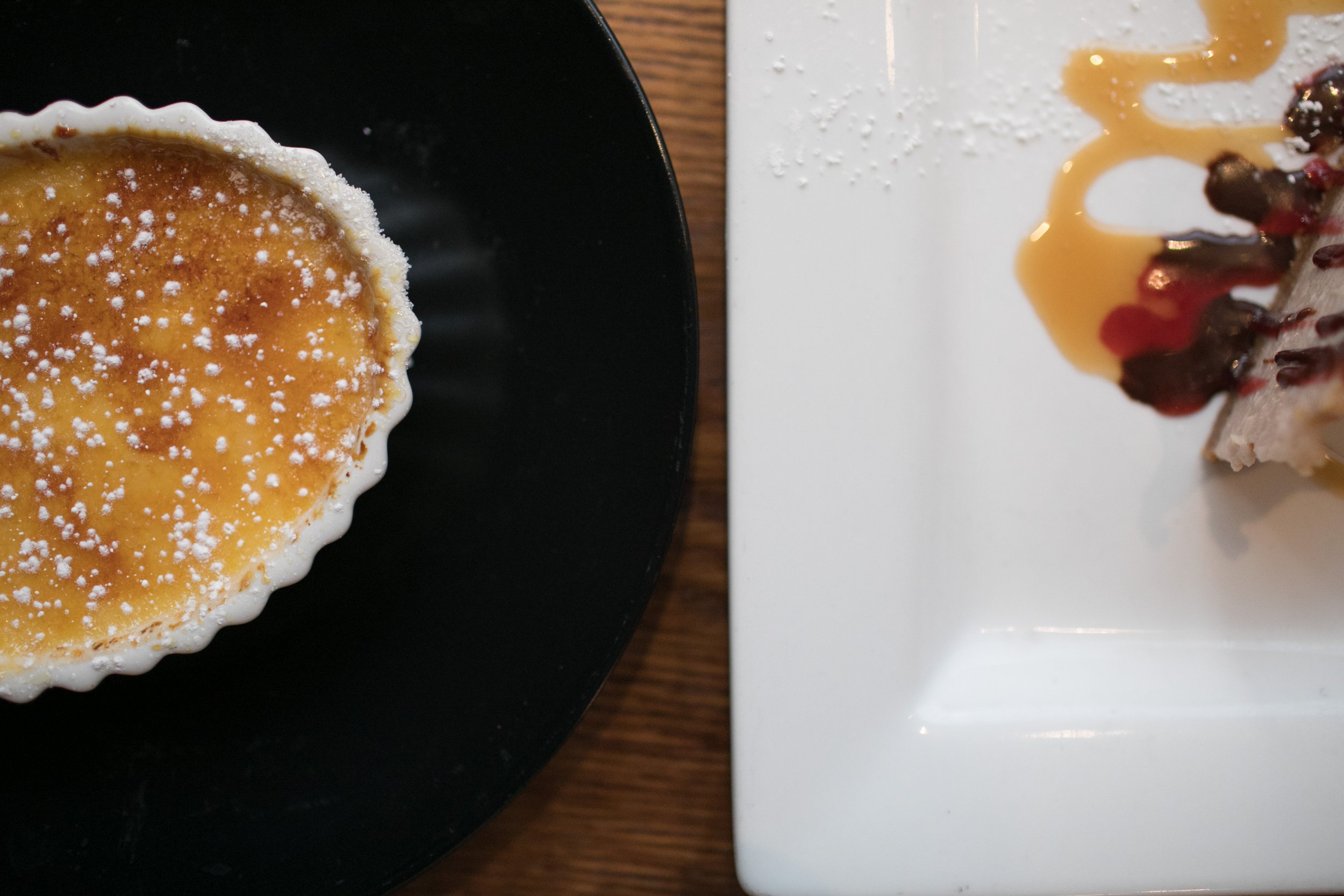 A creme brûlée on a black plate and a cheesecake with caramel sauce and berries on a white plate.