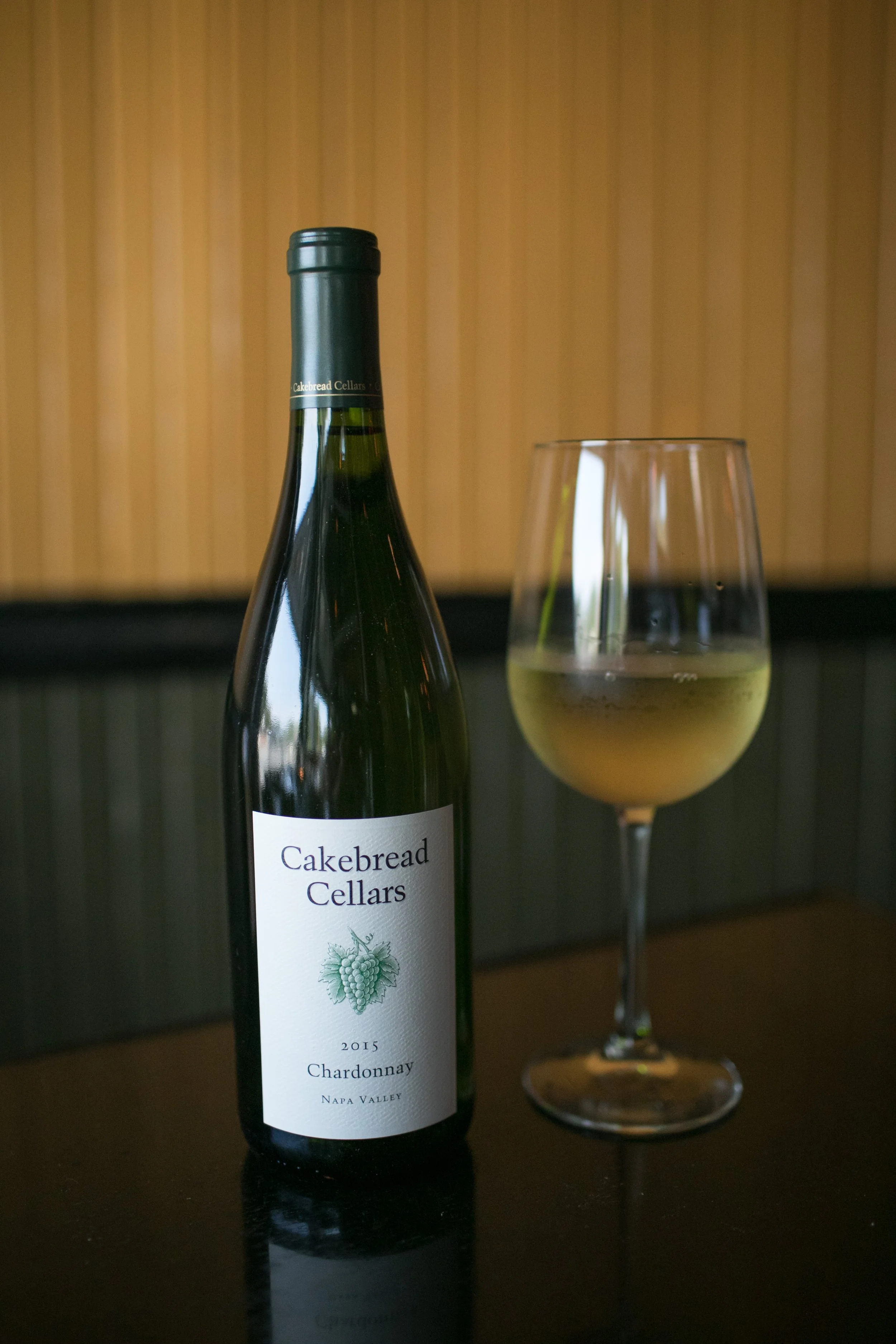 A bottle of Cakebread Cellars 2015 Chardonnay next to a glass of white wine on a black table with a wood-paneled wall background.