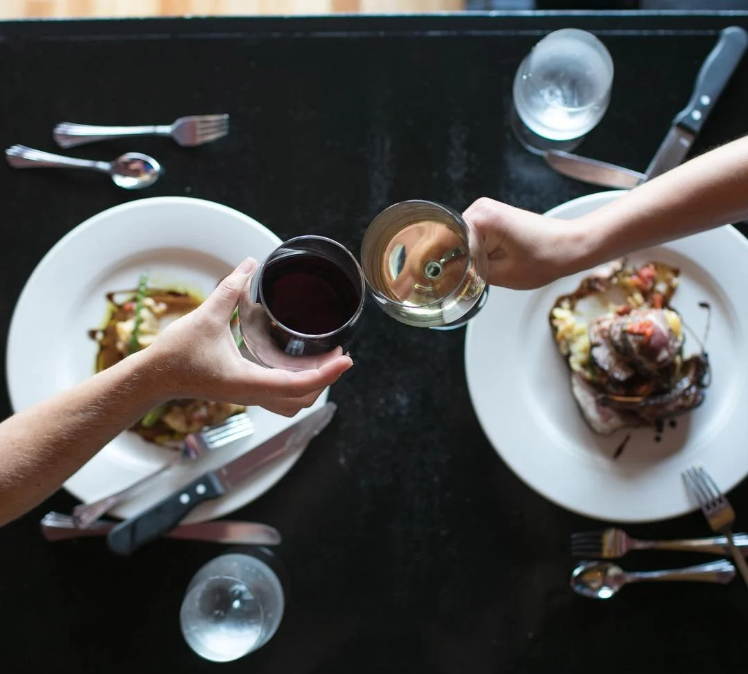Overhead view of two people clinking glasses of red and white wine at a dinner table with plates of food, silverware, and glasses.