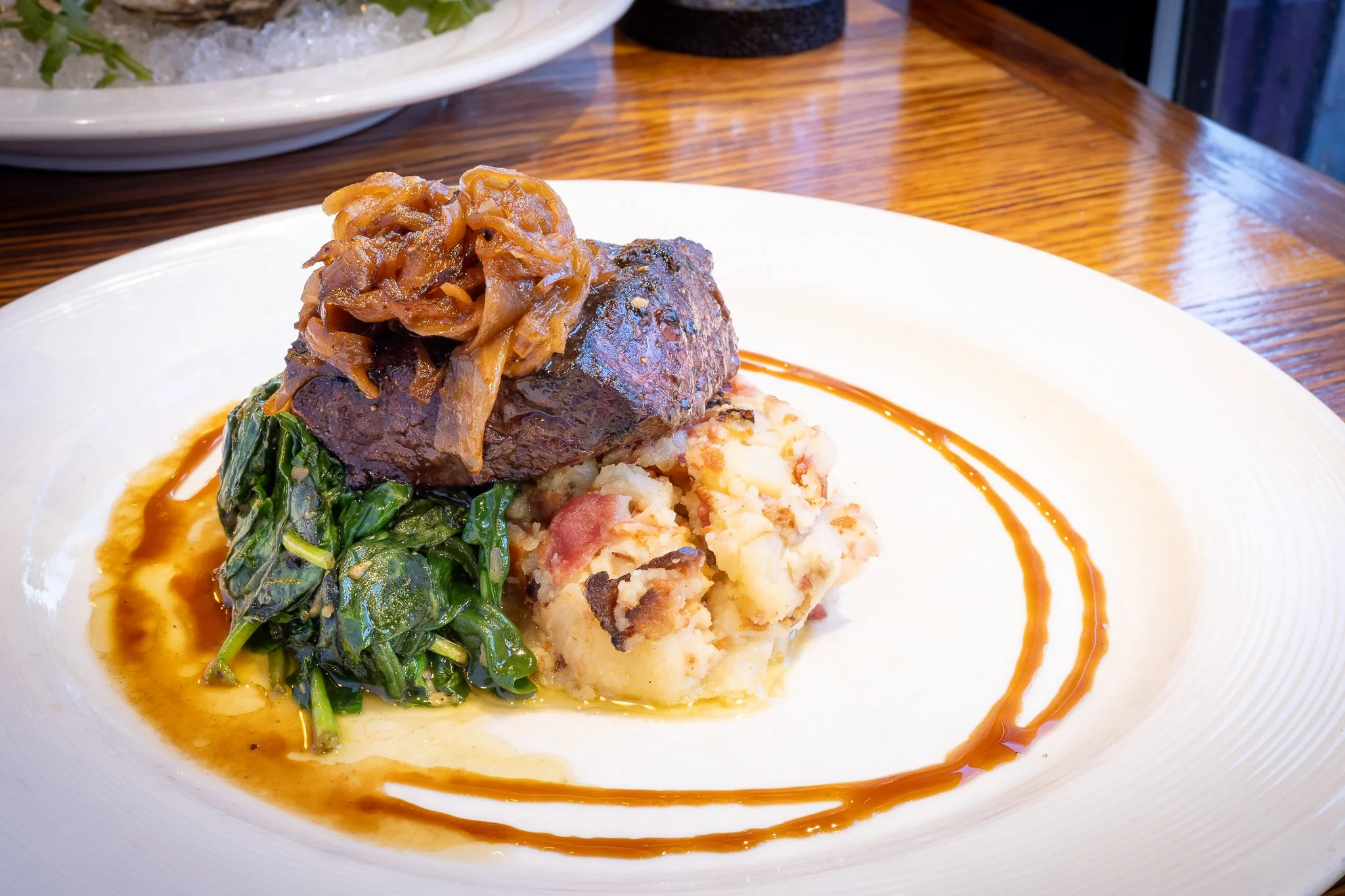 Plate with braised beef topped with caramelized onions, sautéed greens, mashed potatoes with bacon, and a drizzle of sauce on the side
