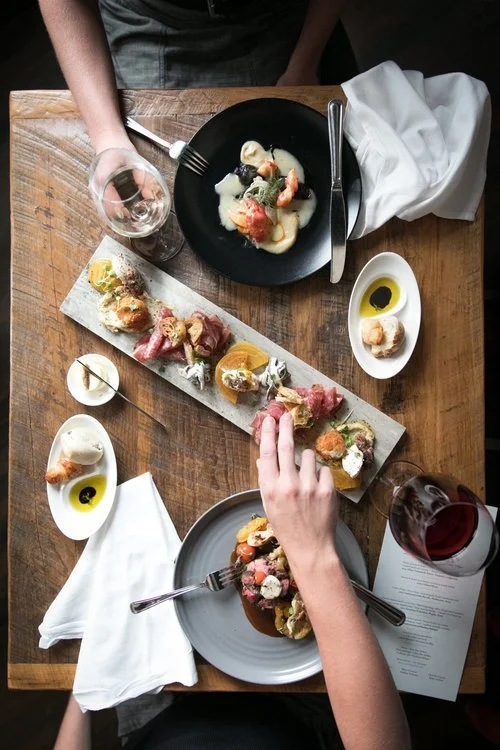 Overhead view of a wooden table with two people dining, featuring various plates of food, wine glasses, and napkins.