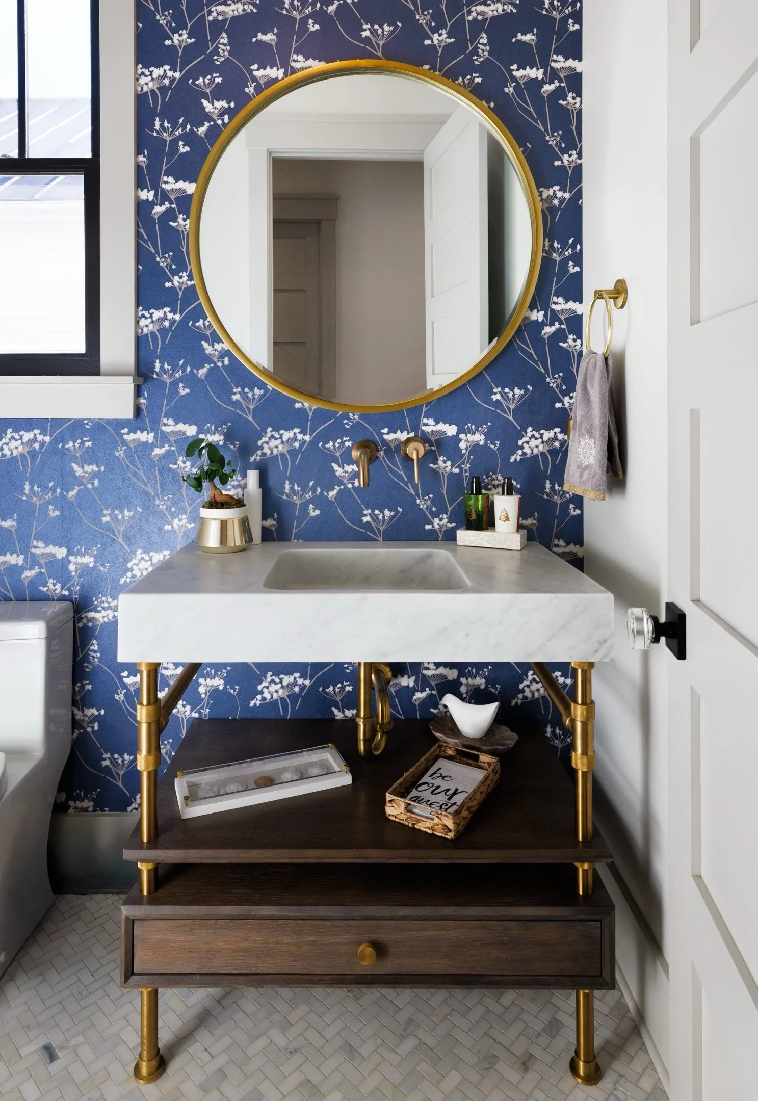 A modern bathroom featuring a white marble sink on a wooden vanity with gold legs, set against a blue floral wallpaper backdrop. A large round gold mirror hangs above the sink, reflecting a small potted plant, toiletries, and a decorative tray on th