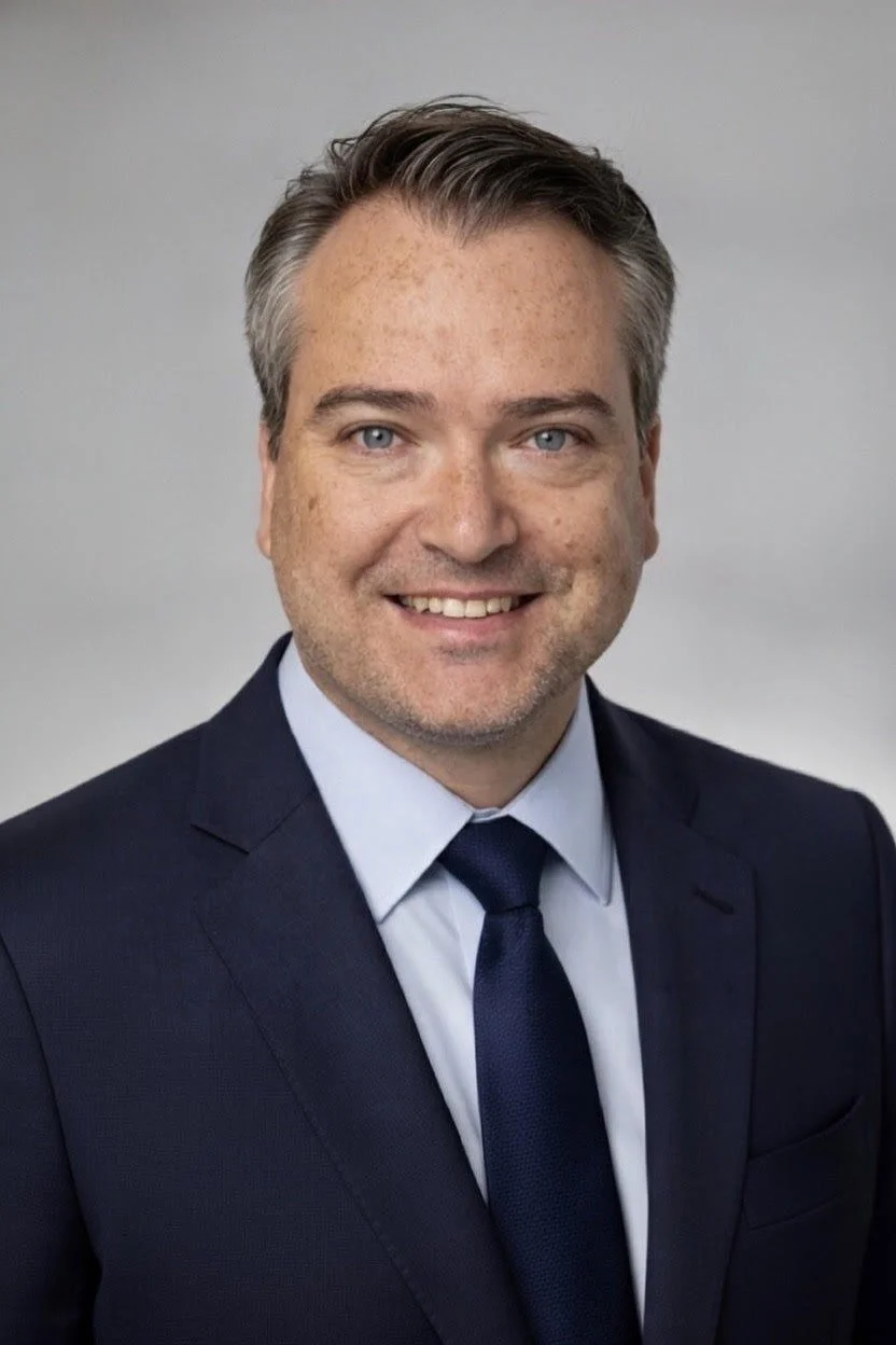 A portrait of a smiling man with light skin, blue eyes, and short, dark hair streaked with gray, wearing a dark suit, light blue shirt, and dark tie, against a light gray background.