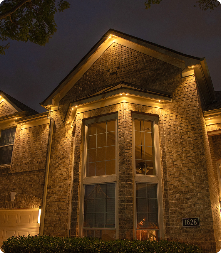 A brick house at night decorated with warm yellow holiday lights outlining the roof and window frame.