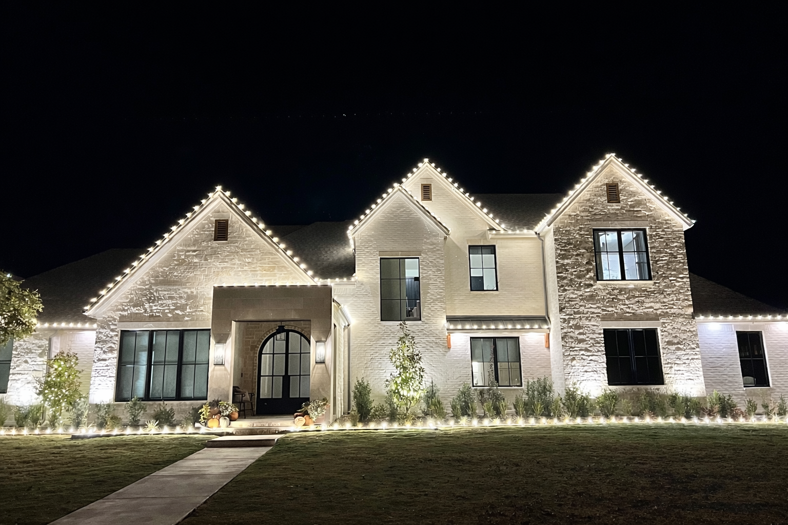 A large, two-story house illuminated with string lights along the roof edges at night, featuring white brick and siding, with a pathway leading to a front door surrounded by potted plants and a landscaped lawn.