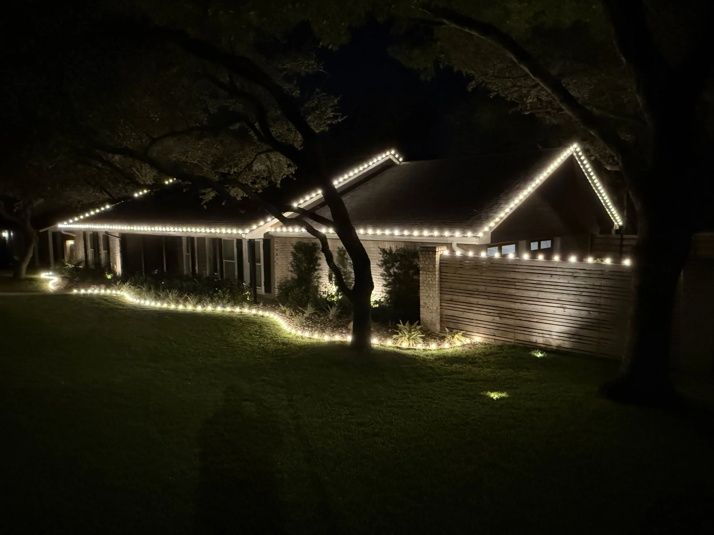A house decorated with white string lights along the roofline, window frames, and garden border at night.