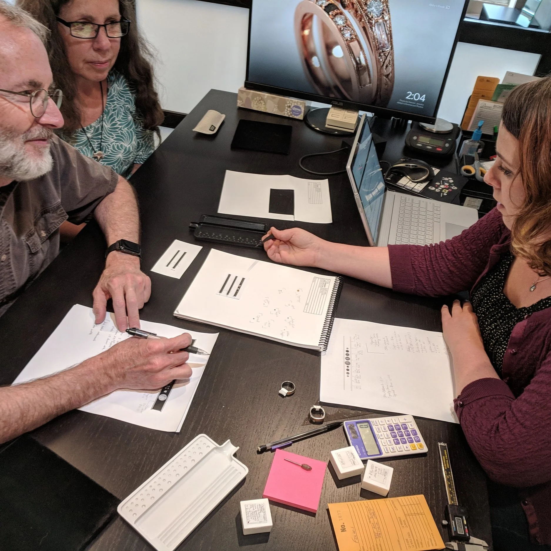 A couple wearing glasses sit across a black desk from their jewelry designer. The designer is sharing sketches and a wax model while one of the customers takes notes.