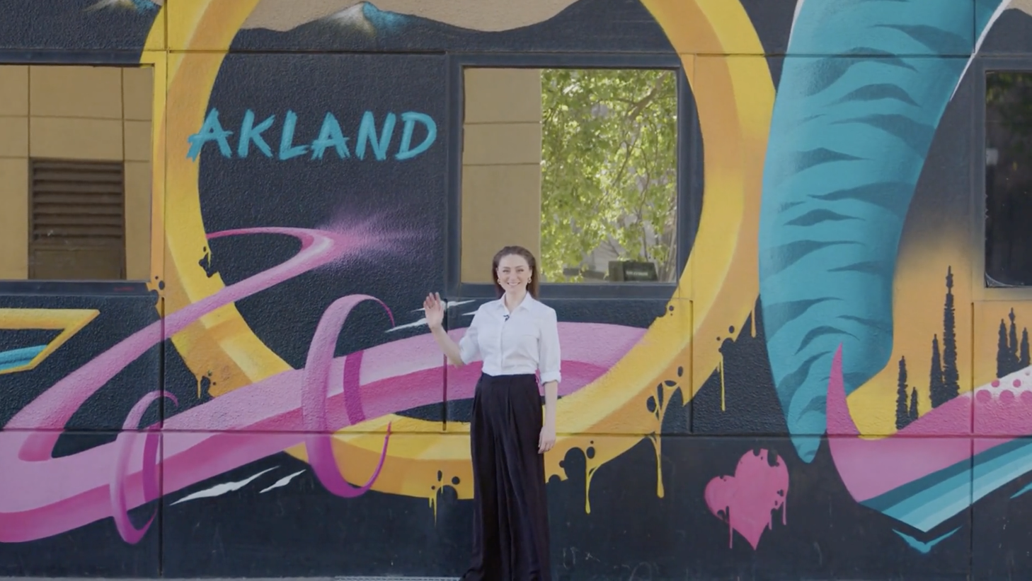A young woman standing in front of a colorful mural on a wall that says "AKLAND" with a pink and purple swirling design, trees, and abstract shapes.