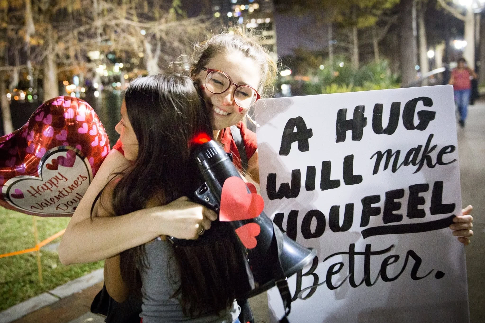 Jamie at a hug march offering a hug to a child while holding a megaphone with red hearts on it. She is also holding a sign that says "a hug will make you feel better" A heart-shaped balloon is in the background.