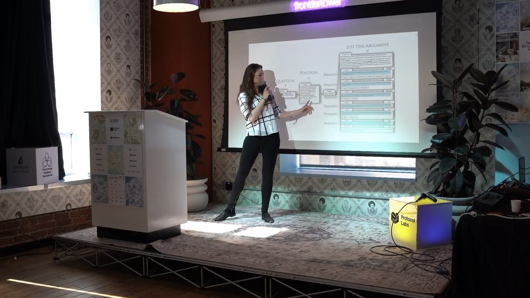 A woman giving a presentation on stage at Protocol Labs, with a large screen displaying a slide titled 'Just One Argument' and various diagrams and text, in a room decorated with patterned wallpaper and plants, and event signage including 'Founder Institute' and 'Furnishing Systems Foundation'.