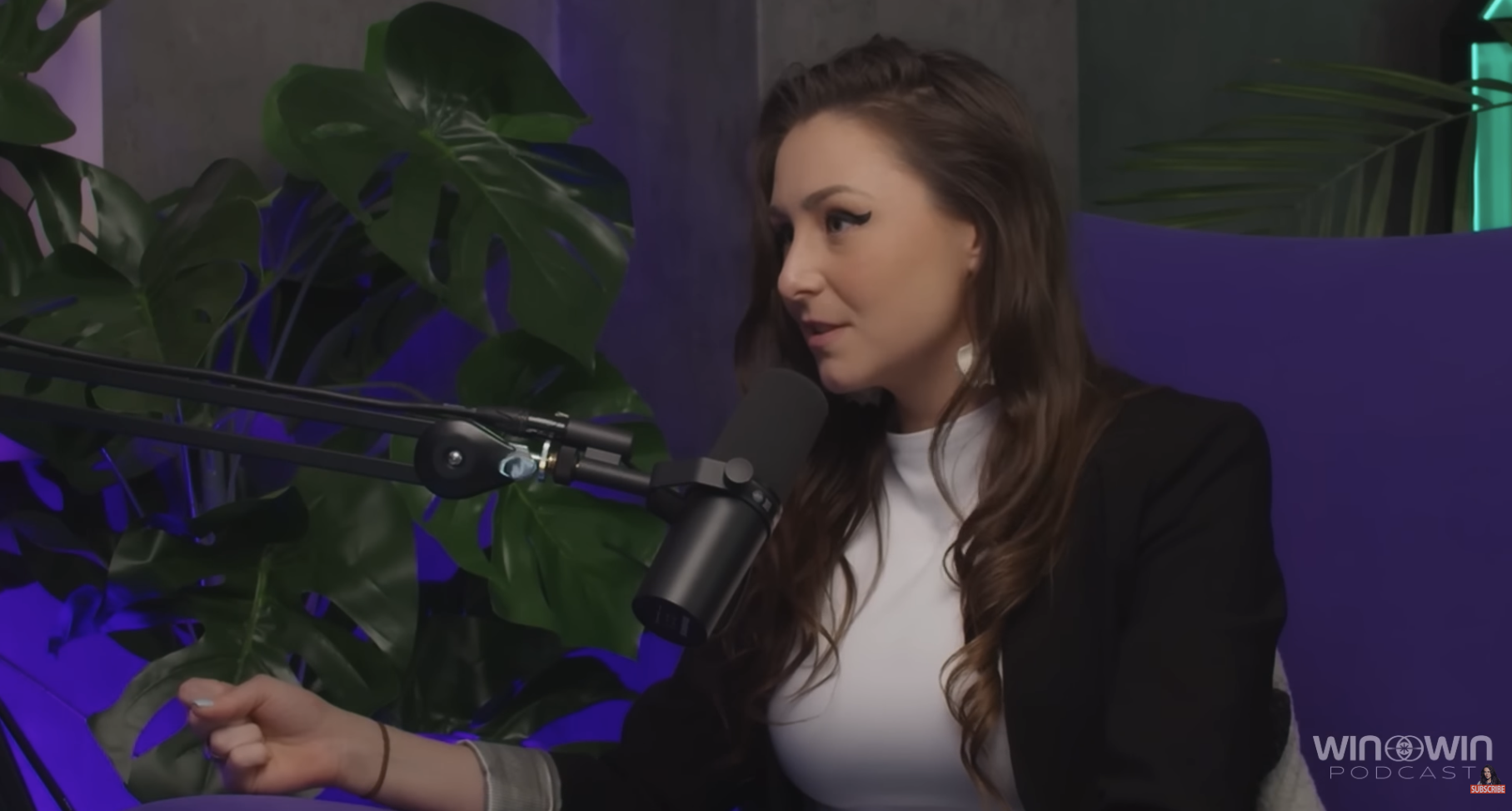 A woman with long brown hair and winged eyeliner speaking into a microphone during a podcast interview, sitting in front of green tropical plants and purple and teal lighting.