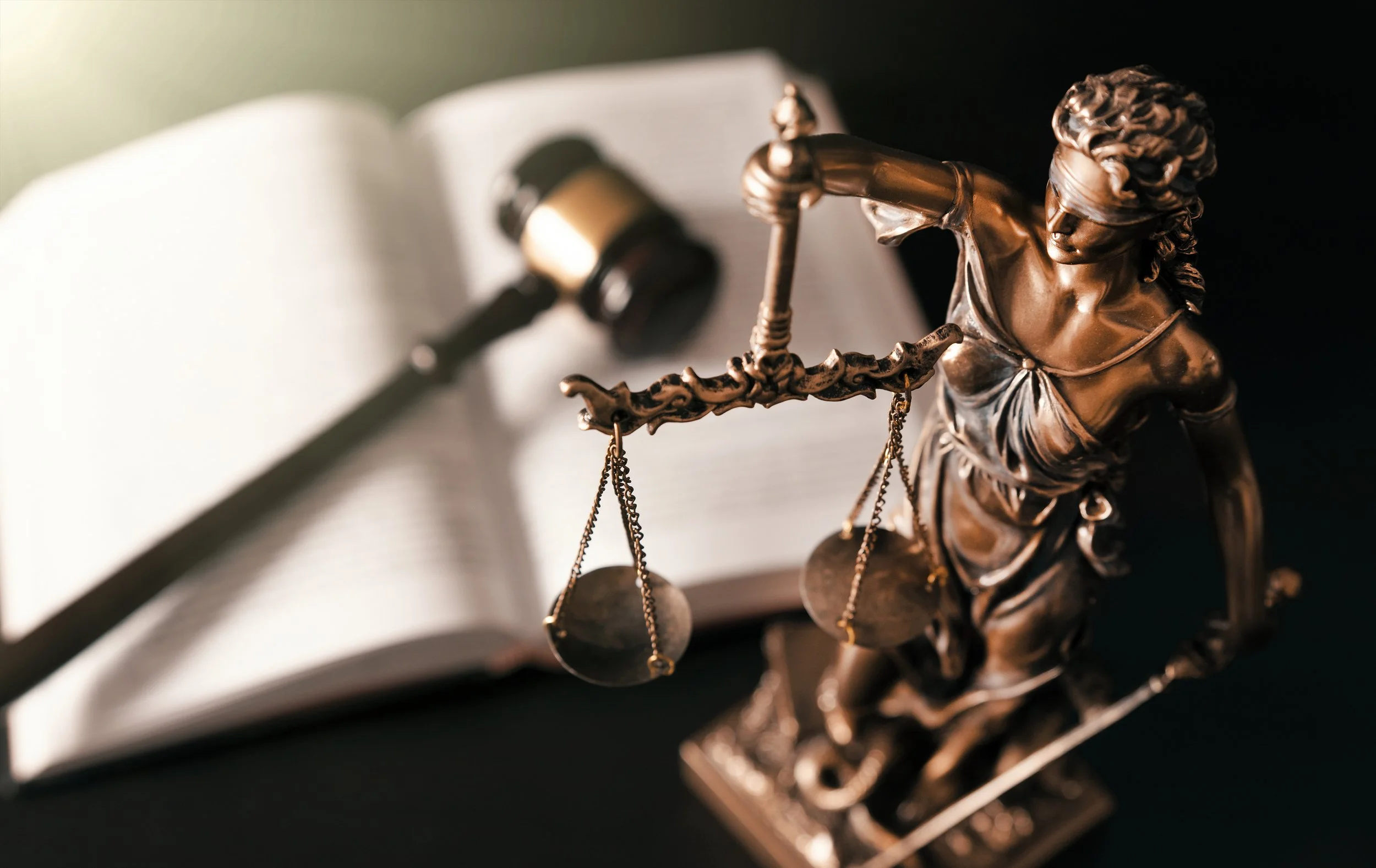 A bronze statue of Lady Justice holding scales, placed on an open law book, with a judge's gavel in the background.