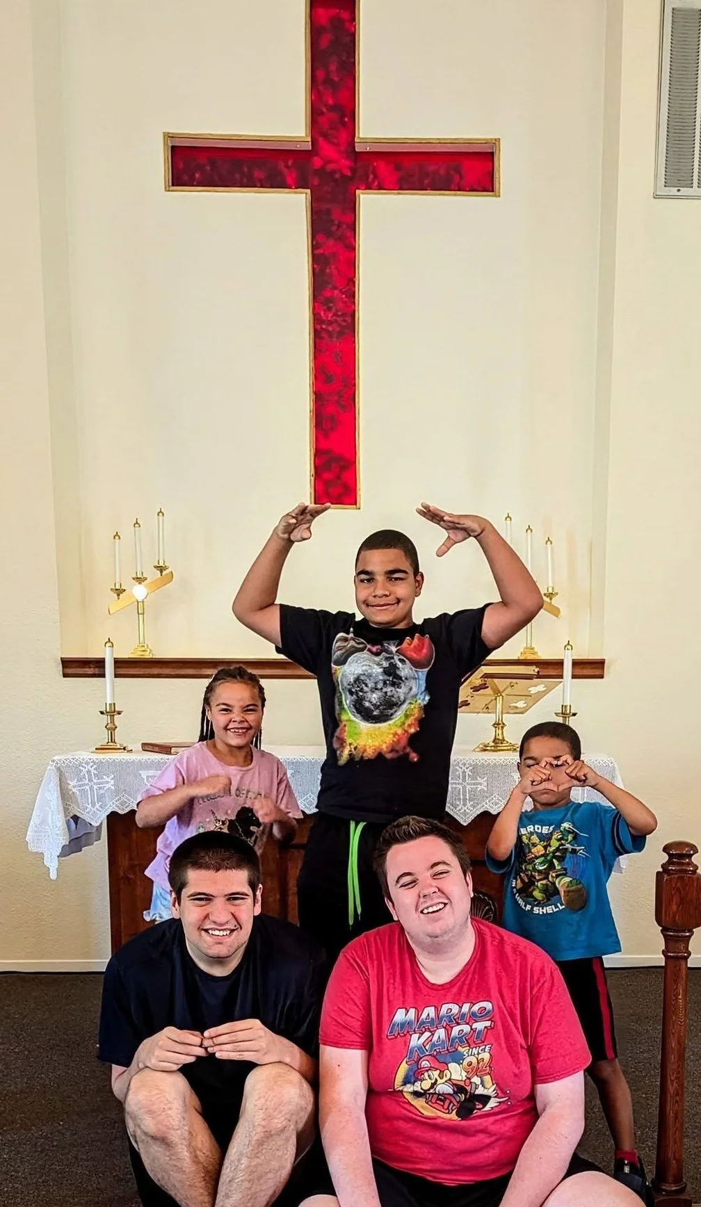 Group of five people posing inside a church with a red cross on the wall behind them. Two young men in front are sitting on the floor, two children and one young man in the back are standing. The children and young man are making playful gestures. An altar with candle holders and a lace cloth is visible in the background.