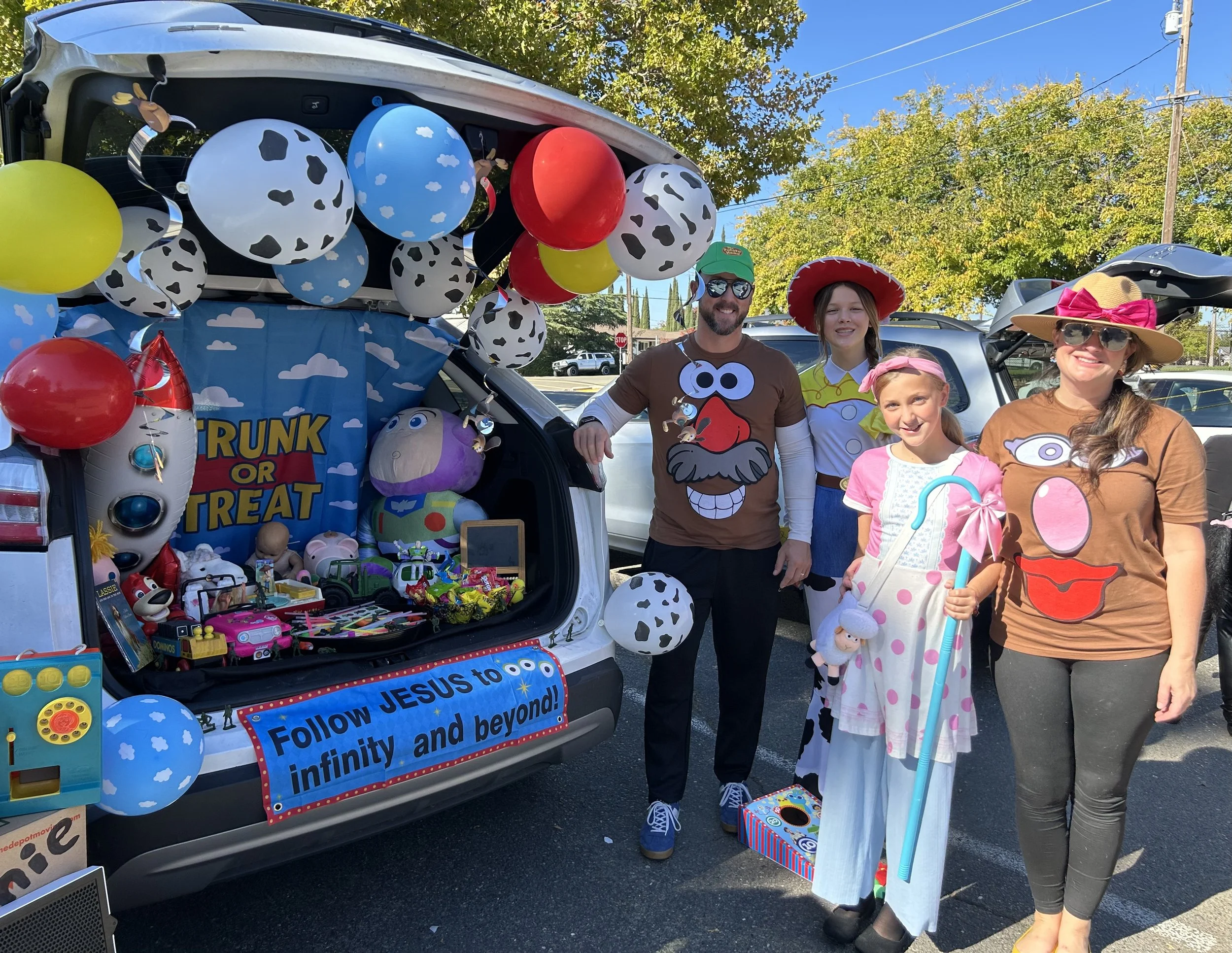 People dressed in costumes standing next to a decorated car with balloons and toys, participating in a festive event outdoors on a sunny day.