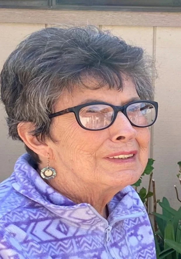 An older woman with short, gray hair and glasses, smiling outdoors, wearing a purple patterned jacket and earrings.