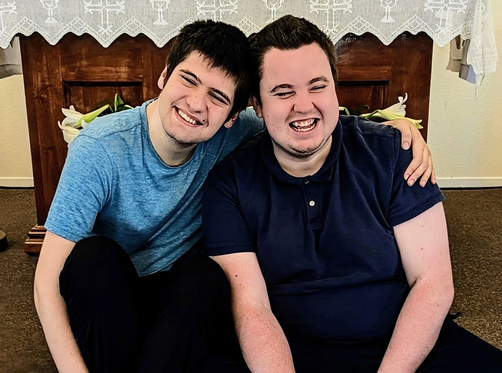 Two young men are sitting on the floor, smiling and laughing, with their arms around each other's shoulders. Behind them, there is a wooden table with white lilies and a lace tablecloth. The background features a beige wall and a carpeted floor.