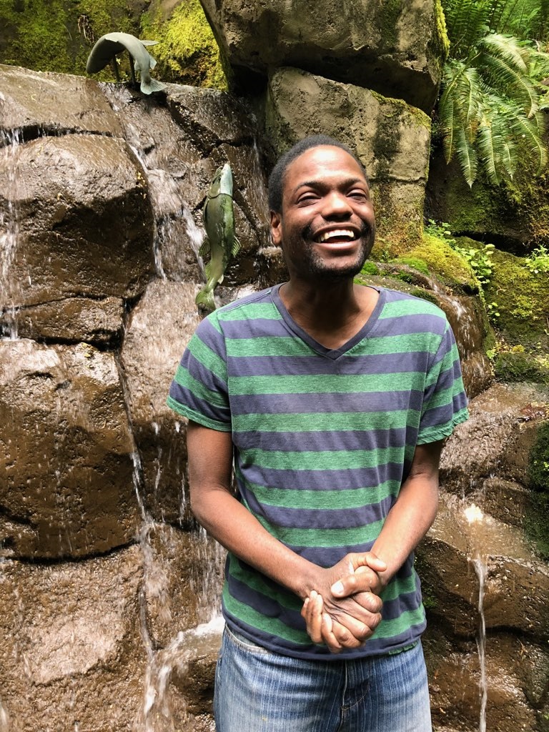 Lawrence smiling widely, clasping his hands together in front of fern and moss covered boulders on a nature walk. Photo courtesy of the artist.