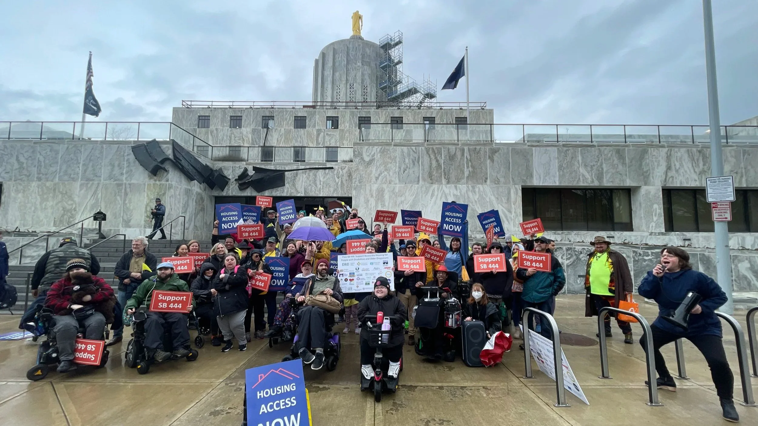 Group of advocates with developmental disabilities and support providers stand on the steps of the Oregon State Capitol Building in Salem, holding signs that say "Housing Access Now" and "Support SB 444"