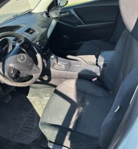 Interior of a Mazda sedan displaying the steering wheel, manual gear shift, handbrake, and dark cloth seats.