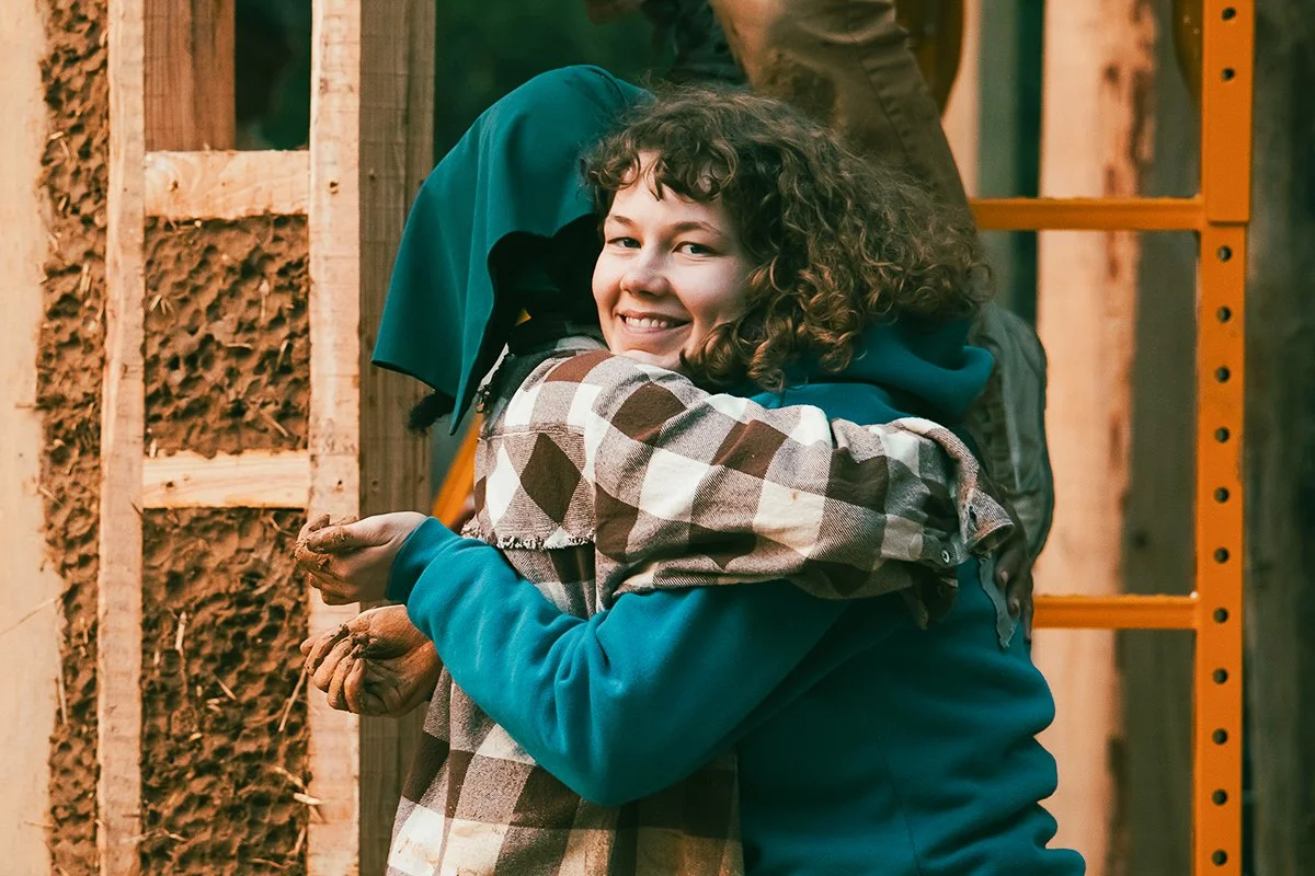A moment of kinship and resilience: Two workshop participants embrace in front of the evolving earthen wall of the Earth Church—hands caked with clay, expressions warm with accomplishment. This hug captures the spirit of community, care, and collabor