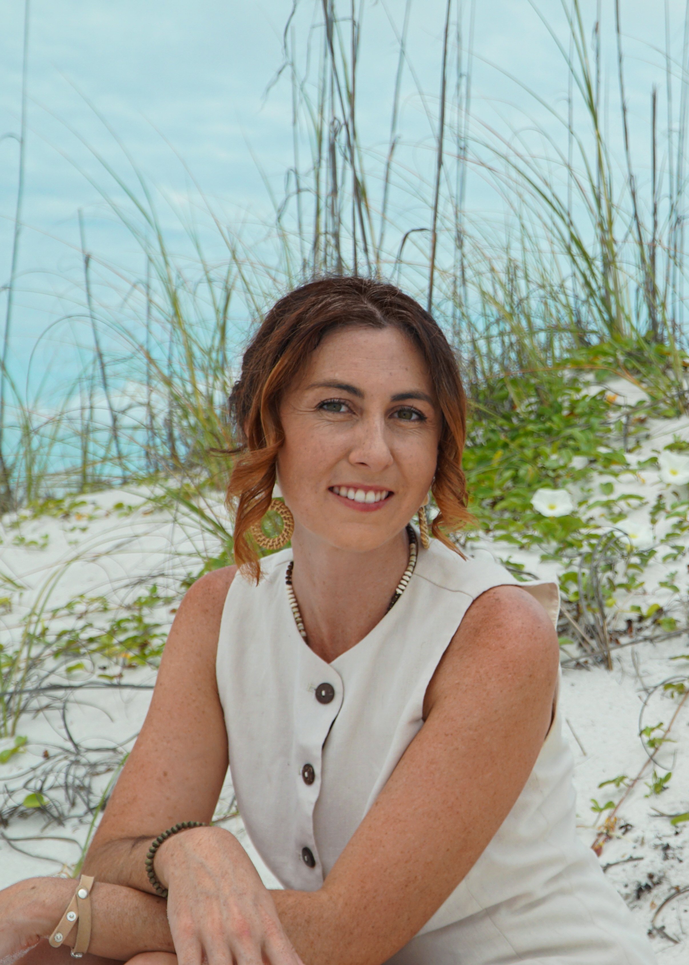 A person with short dark hair smiles at the camera while gently holding the face of a brown and black goat outdoors, with trees and sunlight in the blurred background.
