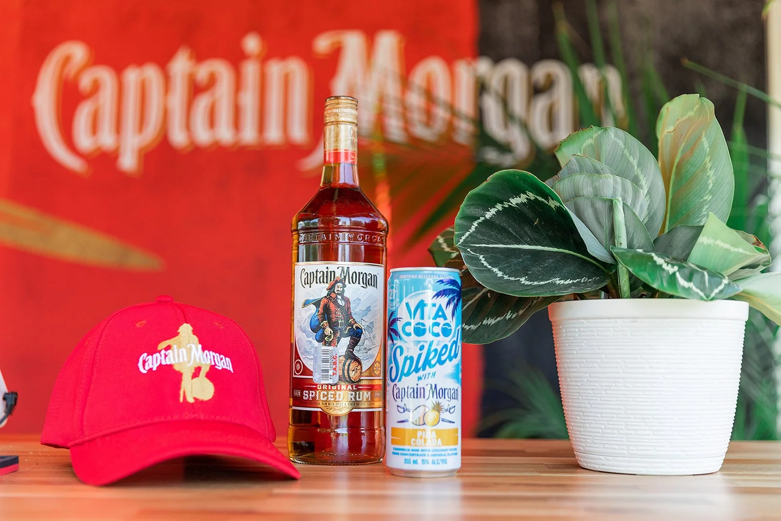 A red Captain Morgan baseball cap, a bottle of Captain Morgan Original Spiced Rum, a can of Captain Morgan Spiked with Pineapple Coconut, and a white pot with green leafy houseplant on wooden surface with Captain Morgan blurred logo background.