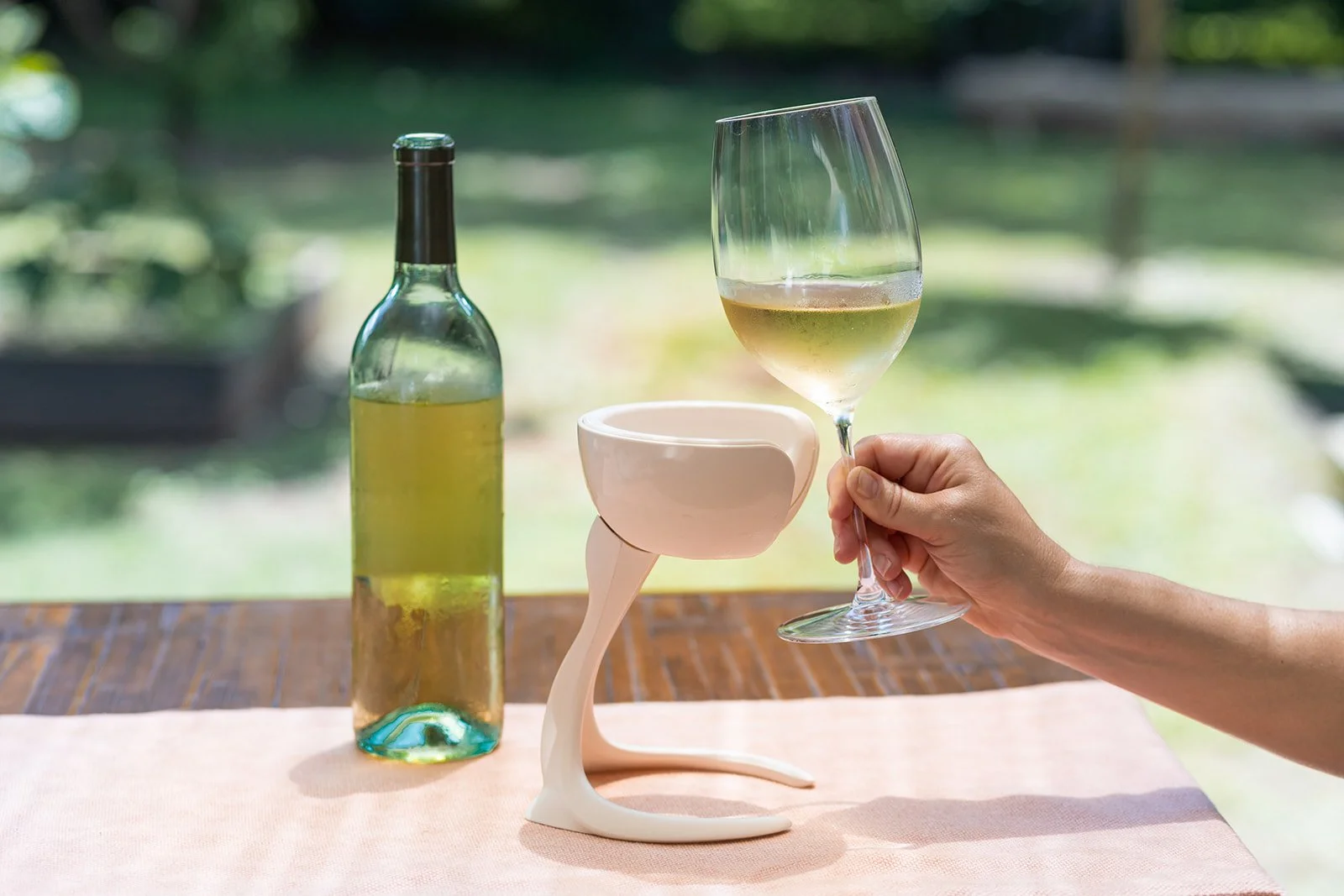 A person’s hand holding a glass of white wine, with a bottle of wine and a white wine glass holder on a table with a pastel pink tablecloth, with a blurred outdoor background.