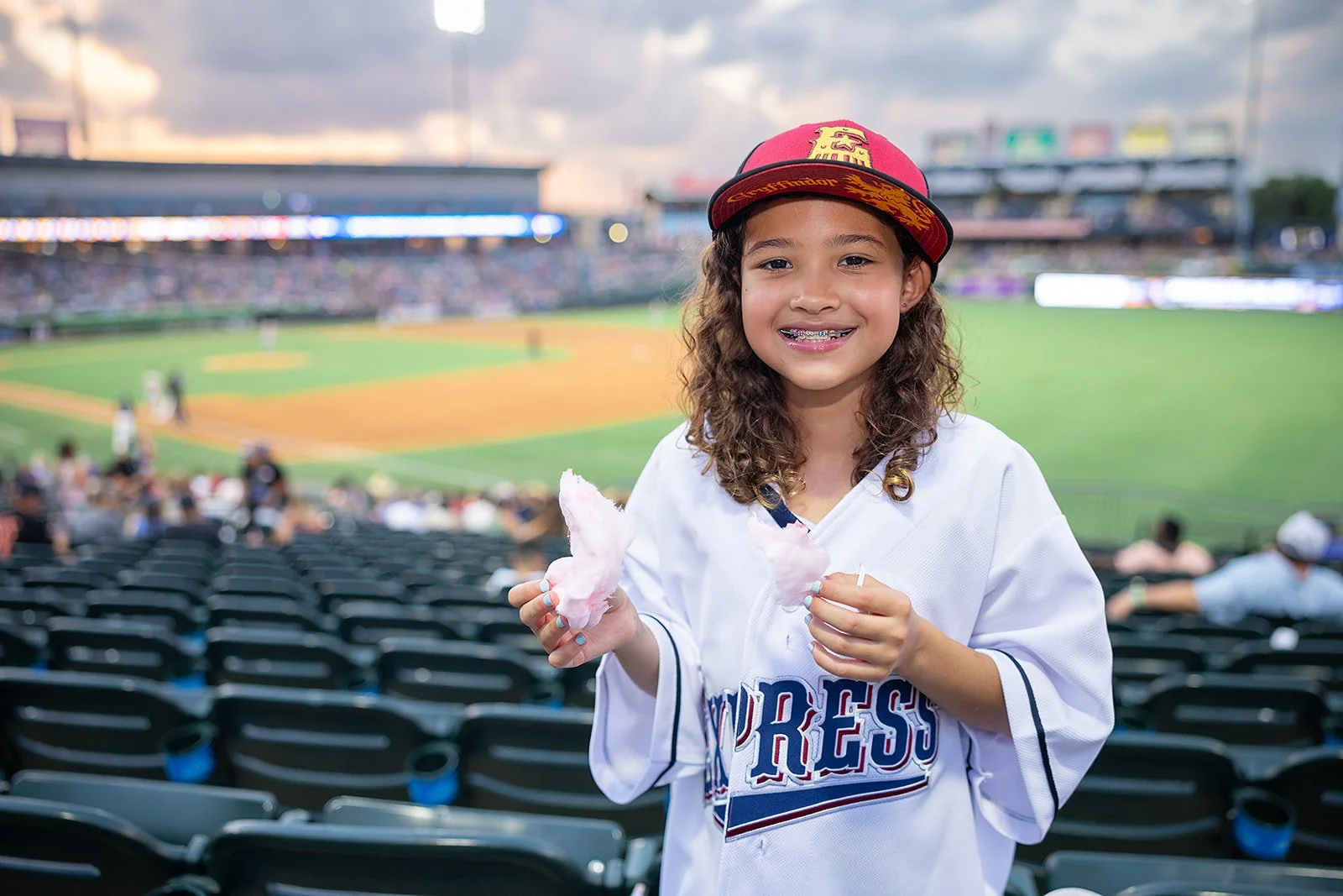 A young girl with curly hair wearing a baseball jersey and a red cap is smiling and holding cotton candy at a baseball stadium, with the field and crowds in the background.