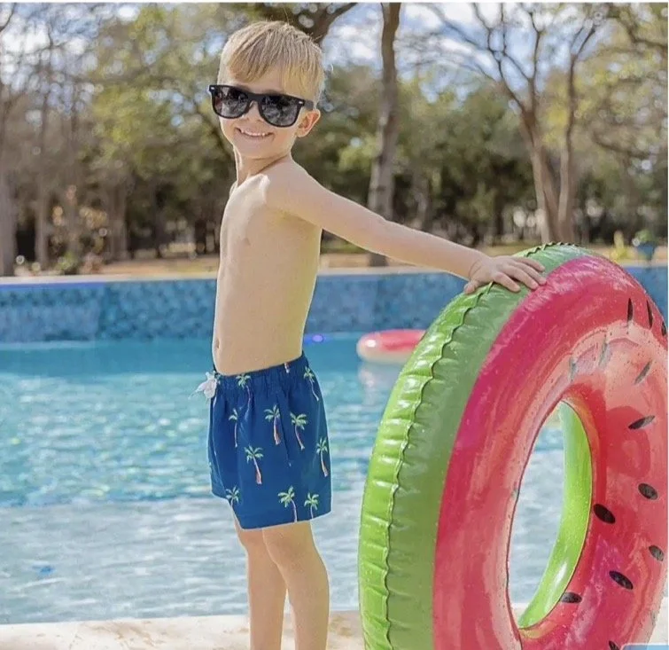  hair wearing sunglasses and blue swim shorts with palm trees, smiling, standing on the edge of a swimming pool, holding a large inflatable float shaped like a watermelon.
