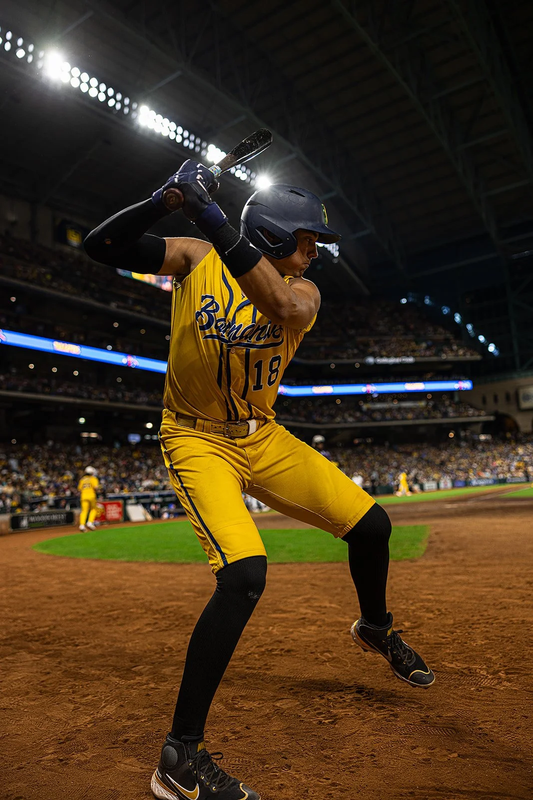 A baseball player in a yellow uniform with the number 18, wearing a helmet and black knee-high socks, is swinging a bat during a game inside a stadium filled with spectators.