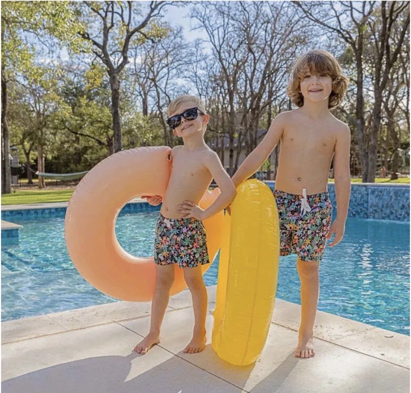  in floral swim trunks standing by a swimming pool with inflatable floats, one with sunglasses and the other smiling, during a sunny day with trees in the background.