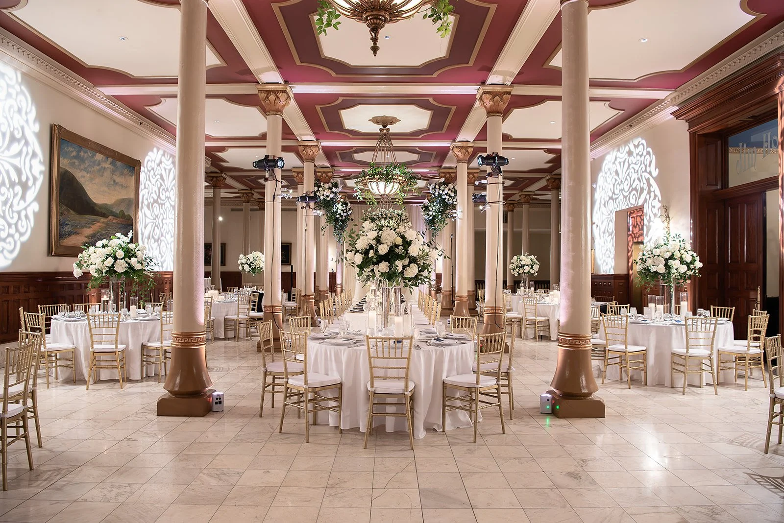 Elegant banquet hall decorated for a wedding or special event with round tables covered in white tablecloths, surrounded by gold chiavari chairs, and adorned with tall floral centerpieces of white flowers and greenery. The ceiling is ornate with pink and cream accents, and there are large columns, chandeliers, and decorative light patterns on the walls.