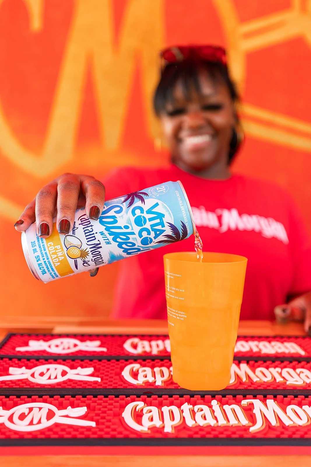 A woman in a red shirt is pouring coconut water into an orange cup at a bar. The background features an orange wall with a large logo, and the bar mat in front says 'Captain Morgan' repeatedly.