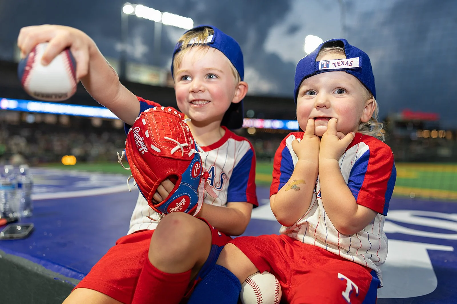 Two young children wearing Texas Rangers baseball uniforms and caps sitting in a baseball stadium, one holding a baseball glove and ball, smiling and posing for the camera.