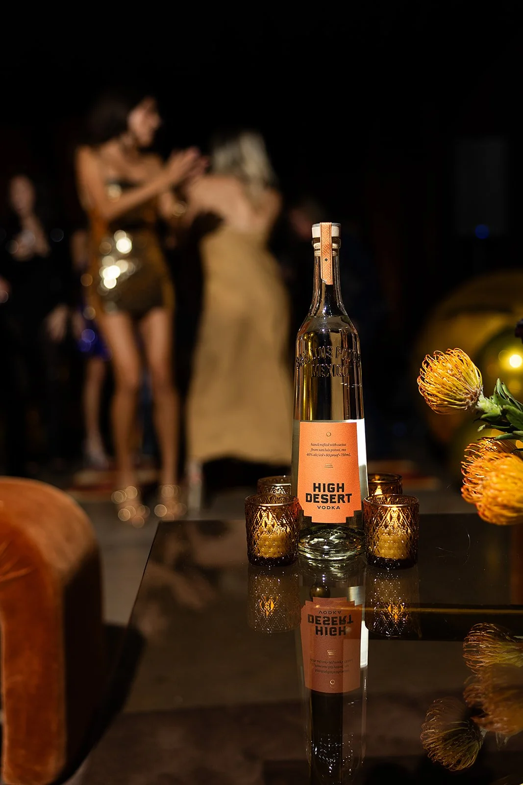 A bottle of High Desert vodka placed on a reflective black table, surrounded by small candle holders. Blurred people in golden attire are in the background at a dimly lit social gathering.