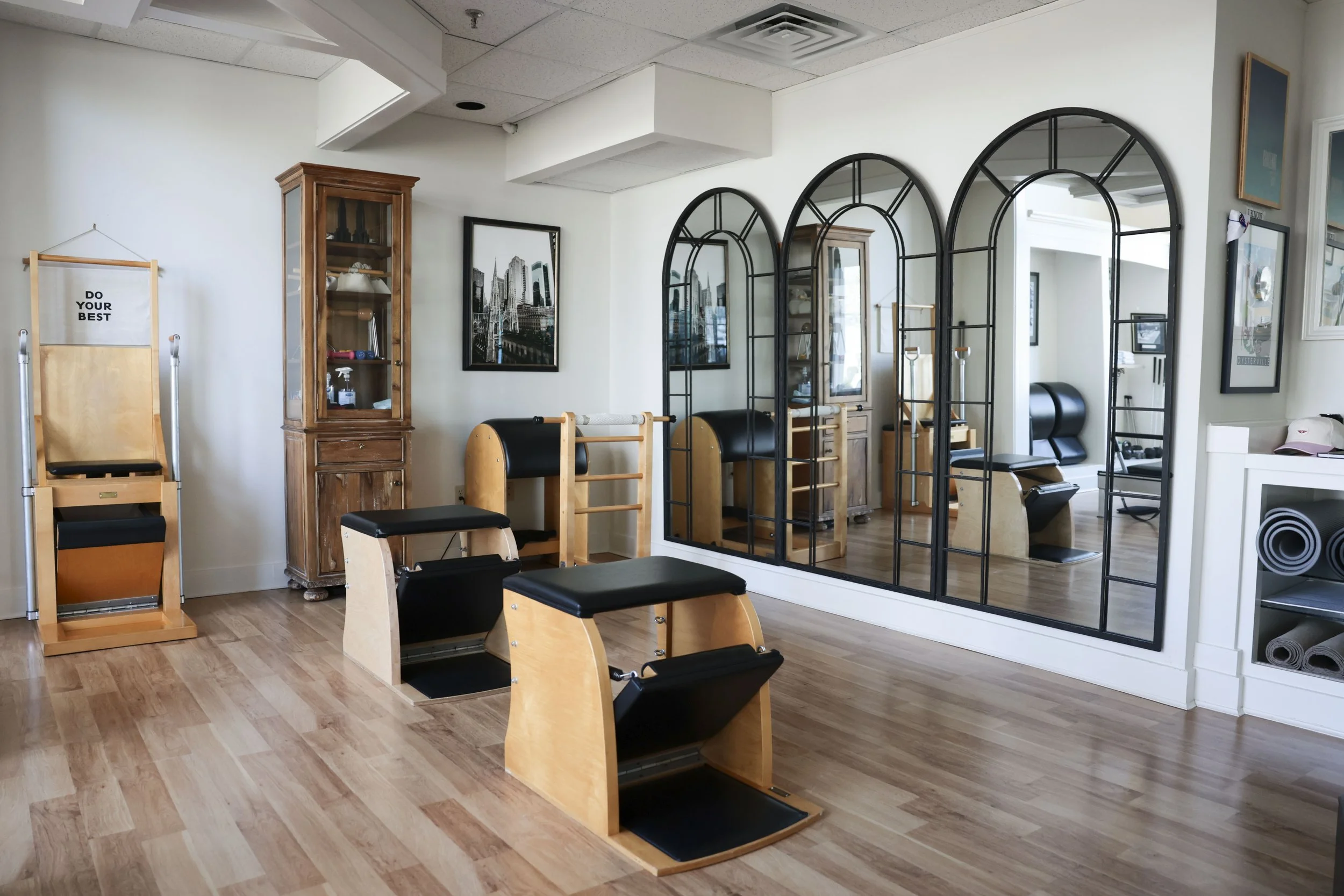 A Pilates studio with wooden and black equipment, large arched mirrors on the wall, framed artwork, a wooden cabinet, and rolled-up yoga mats on a shelf.