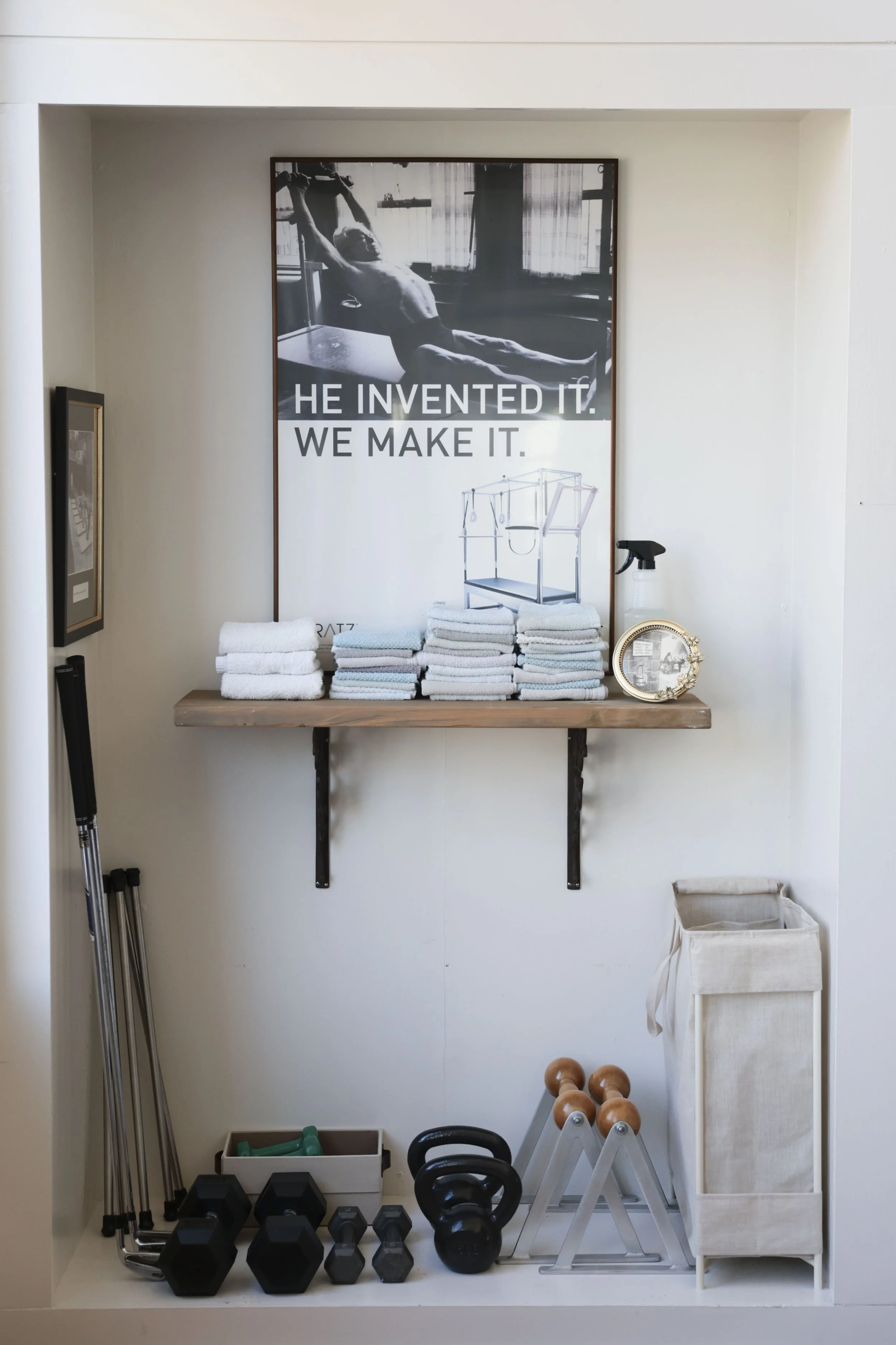 A display of organized fitness and gym equipment in a small corner with a shelf holding folded towels, a spray bottle, and a decorative frame; below are kettlebells, dumbbells, and exercise accessories; a large motivational poster on the wall feature