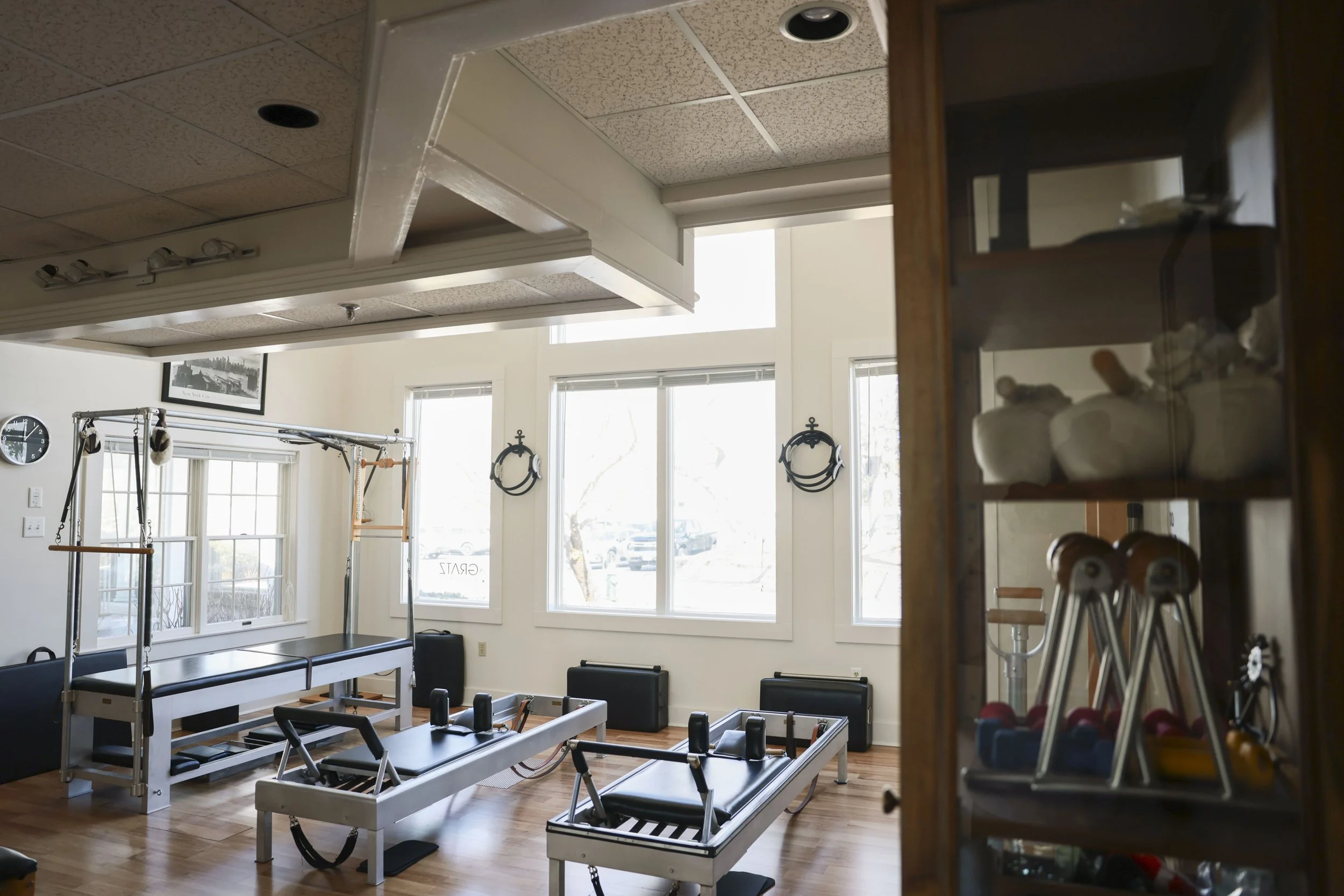Pilates studio with reformer machines, large windows, wall decor, and a wooden cabinet with exercise equipment.