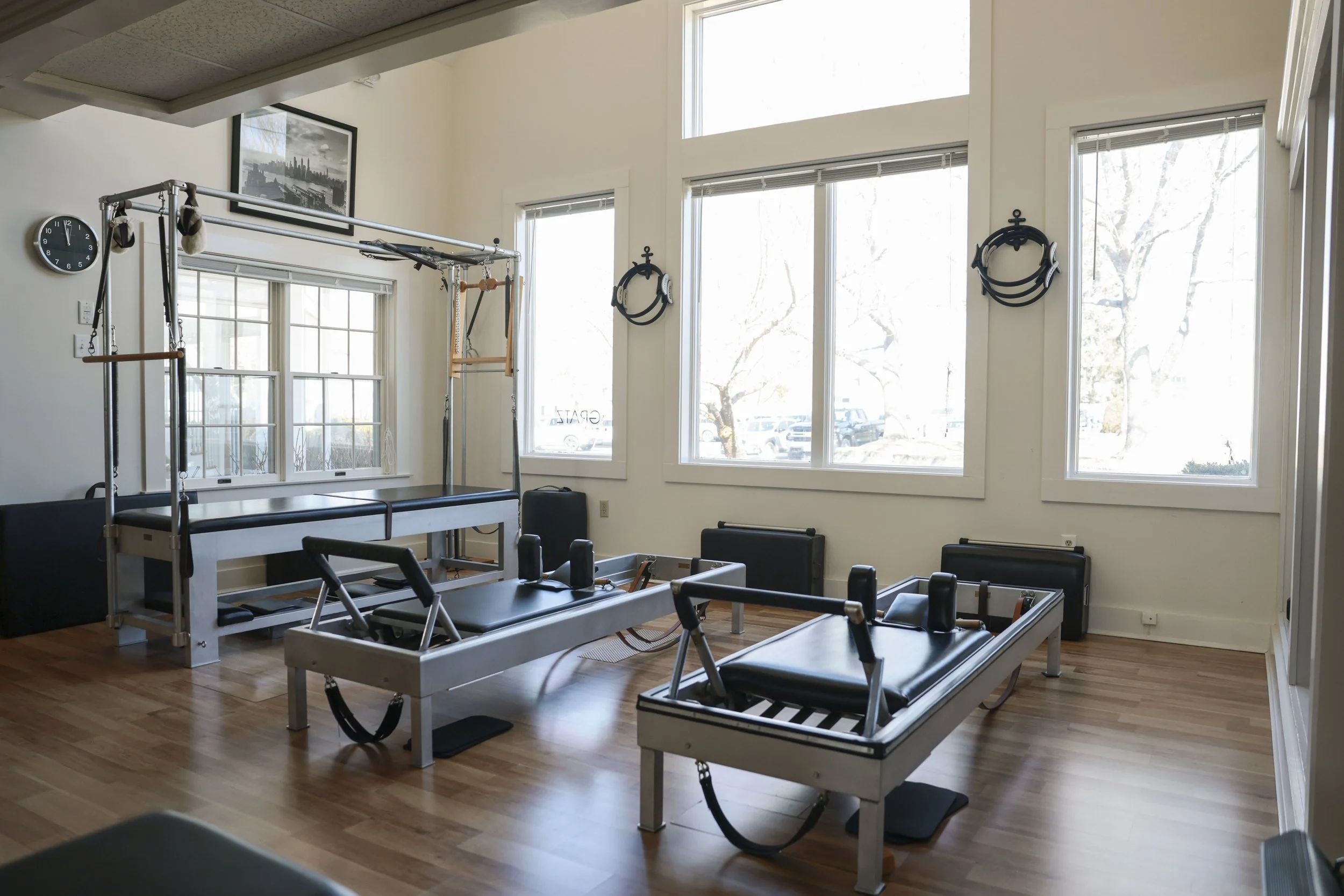 Fitness studio with Pilates reformers, chairs, and aerial equipment. Large windows let in natural light, with a clock and framed picture on the wall.