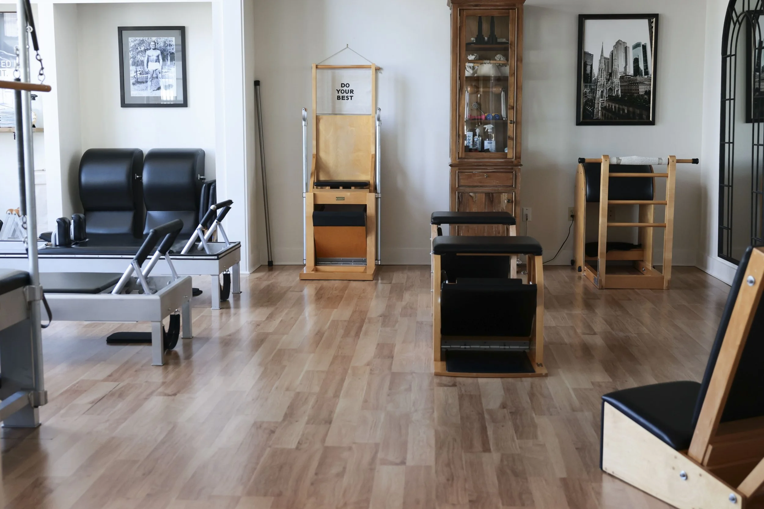 Pilates equipment including reformers, chairs, and a stability barrel in a room with wooden floors, framed photographs on the walls, and a wooden cabinet with glass doors.