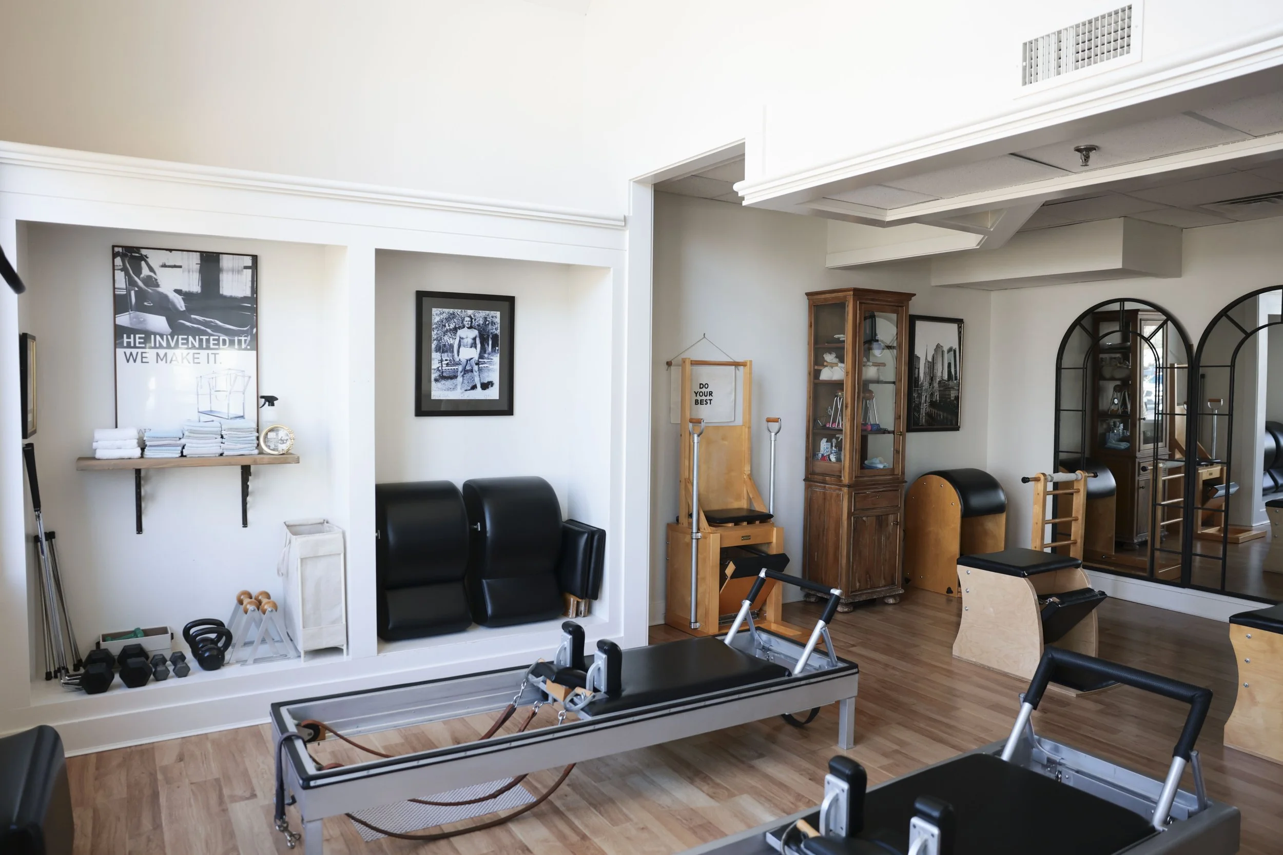 A Pilates or fitness studio with equipment, chairs, and shelves, including wall art and framed photos, in a well-lit room with wooden floors.